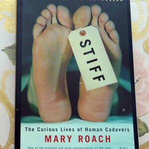 May include: A book cover with the title "STIFF" on a tag attached to a pair of feet. The book is titled "The Curious Lives of Human Cadavers" by Mary Roach. The cover also includes the text "NEW YORK TIMES BESTSELLER".
