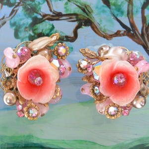 May include: Vintage clip-on earrings with a floral design. Each earring features a large coral flower with a central crystal, surrounded by pink beads, pearls, and gold filigree. The background is a painted landscape scene.