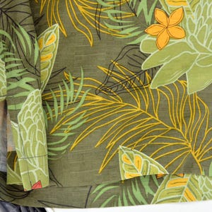 K&ouml;nnte beinhalten: Nahaufnahme eines gr&uuml;nen Stoffes mit tropischem Blumenmuster. Das Design zeigt gro&szlig;e wei&szlig;e und gr&uuml;ne Blumen, gelbe und gr&uuml;ne Palmenbl&auml;tter sowie orange und rote Bl&uuml;ten. Der Stoff ist gefaltet und gibt die Innenseite frei.