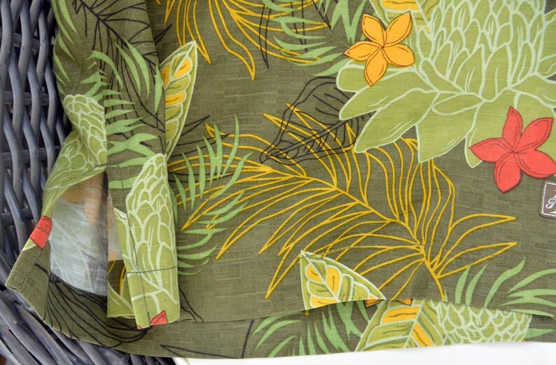 K&ouml;nnte beinhalten: Nahaufnahme eines gr&uuml;nen Stoffes mit tropischem Blumenmuster. Das Design zeigt gro&szlig;e wei&szlig;e und gr&uuml;ne Blumen, gelbe und gr&uuml;ne Palmenbl&auml;tter sowie orange und rote Bl&uuml;ten. Der Stoff ist gefaltet und gibt die Innenseite frei.