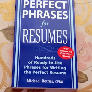May include: A blue book titled "PERFECT PHRASES for RESUMES" with white text. It includes the text "Hundreds of Ready-to-Use Phrases for Writing the Perfect Resume" and a red sticker that says "Key Terms to Get You Hired".