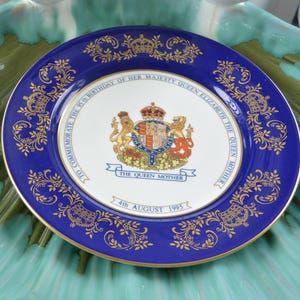 May include: A commemorative plate with a blue rim and gold filigree. The center features a royal crest and the text "TO COMMEMORATE THE 95th BIRTHDAY OF HER MAJESTY QUEEN ELIZABETH THE QUEEN MOTHER 4th AUGUST 1995".