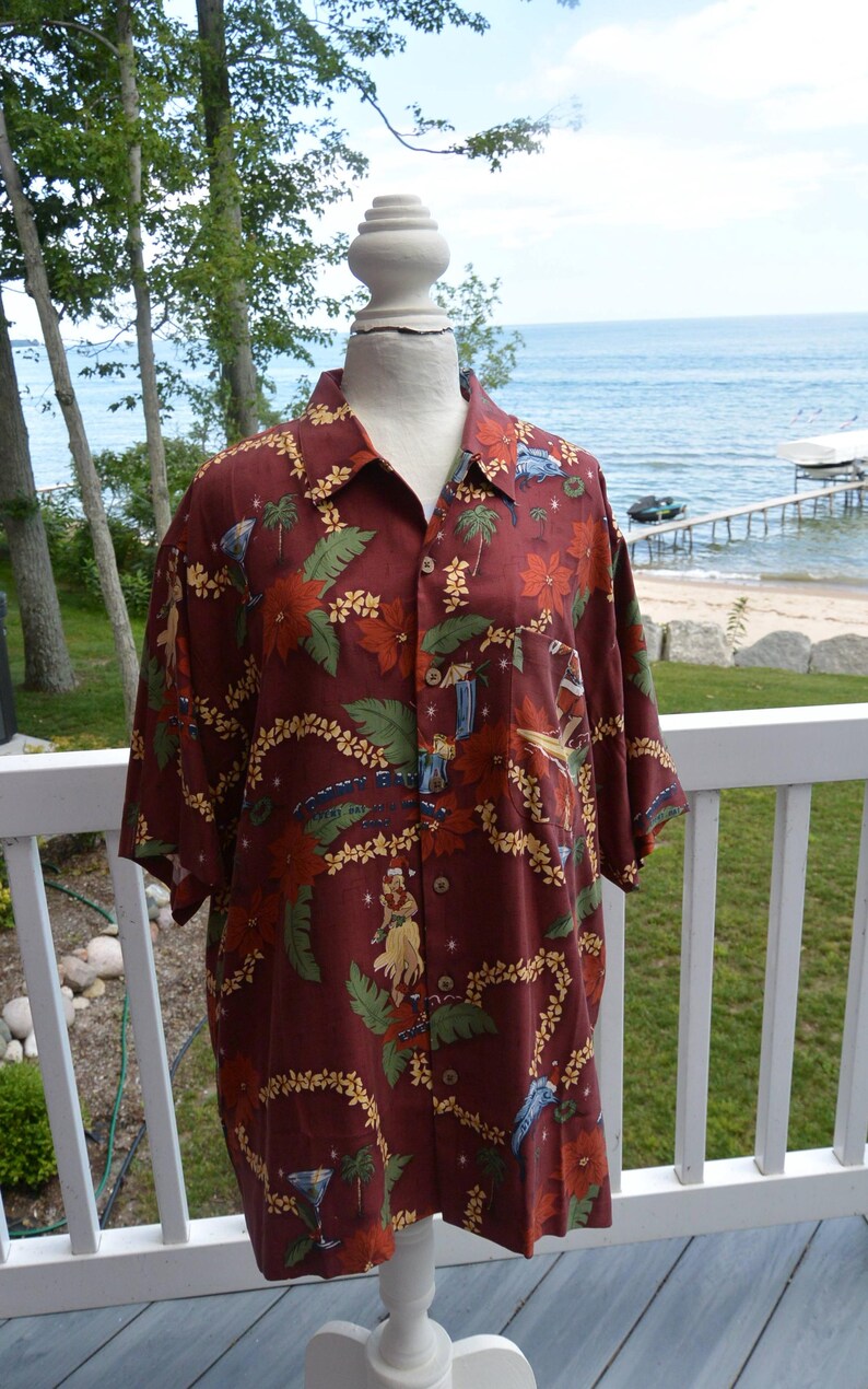 May include: Burgundy short-sleeve button-down shirt with a tropical floral and figure print. The shirt features a classic collar, a chest pocket, and a relaxed fit. The design includes palm trees, cocktails, and hula dancers.