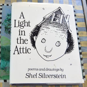 May include: A hardcover book titled "A Light in the Attic" by Shel Silverstein. The cover features a black and white illustration of a face with a house on top of the head. The book is white with black text.