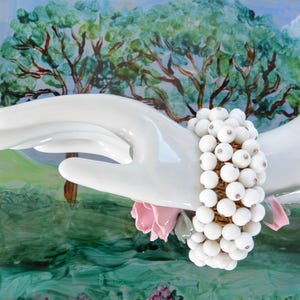 May include: A white beaded bracelet with a gold clasp. The bracelet is on a porcelain hand with a pink flower.