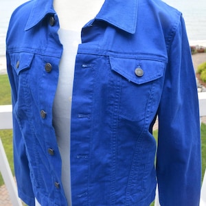 May include: A vibrant royal blue denim jacket featuring a classic collar, long sleeves, and a front button-down closure. The jacket includes two chest pockets with buttoned flaps and vertical seam detailing, displayed on a white mannequin torso.