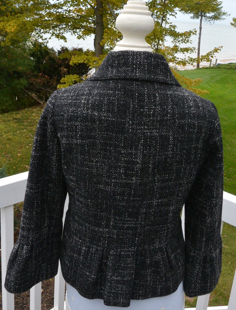 May include: Black and white tweed jacket with a fitted silhouette and a ruffled hem. The jacket has a collar and long sleeves.