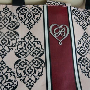 May include: Tote bag featuring a cream-coloured background with a black damask pattern. A central burgundy stripe with a silver heart-shaped emblem, featuring the letter "B".