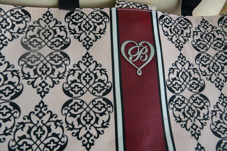 May include: Tote bag featuring a cream-coloured background with a black damask pattern. A central burgundy stripe with a silver heart-shaped emblem, featuring the letter "B".