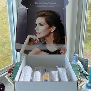May include: An open white box containing beauty products, with a picture of Cindy Crawford on the inside lid. The box is labeled "Meaningful Beauty By Design". The products are in clear plastic packaging.