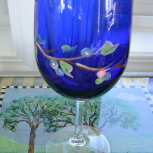 May include: A cobalt blue wine glass with a clear stem and base. The glass is decorated with a painted design of a brown branch with green leaves and pink and white berries. The glass is on a painted surface.