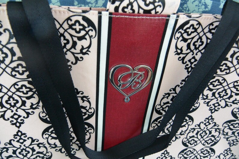 May include: A tote bag featuring a pink and black floral design. The bag has a central red stripe with a silver heart-shaped emblem. Black straps are attached to the top of the bag.