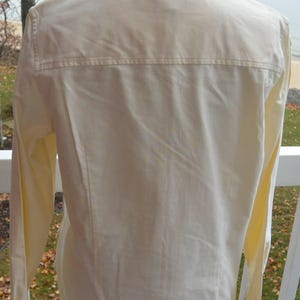 Puede incluir: Vista trasera de una camisa abotonada de color amarillo p&aacute;lido. La camisa tiene mangas largas, cuello y un detalle de canes&uacute; en la parte superior de la espalda. La tela parece ser un material ligero.