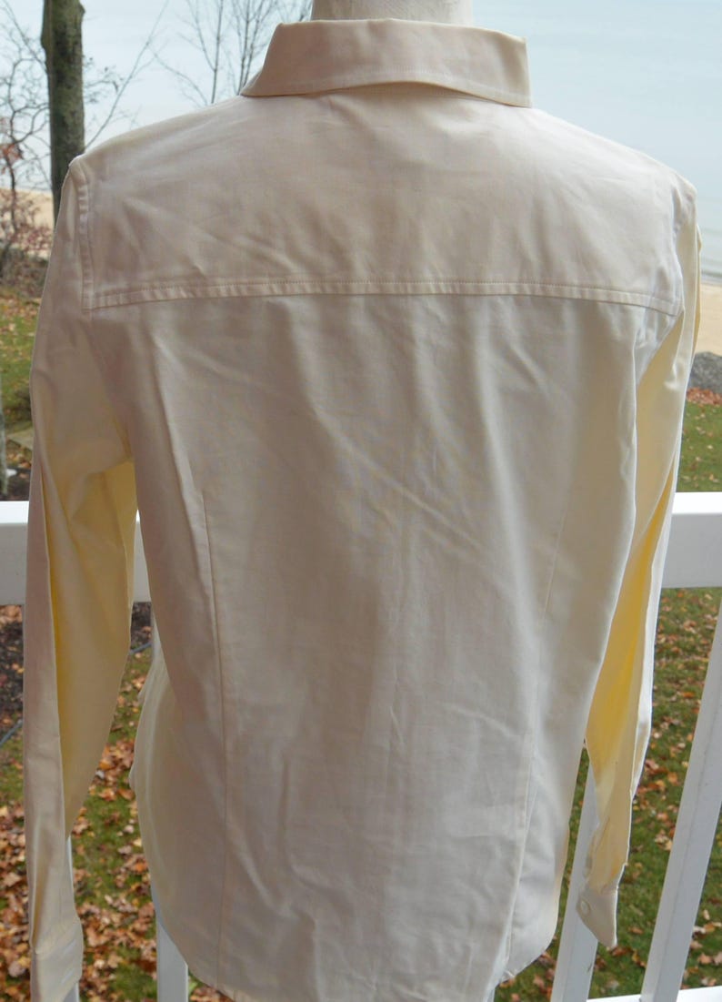 Puede incluir: Vista trasera de una camisa abotonada de color amarillo p&aacute;lido. La camisa tiene mangas largas, cuello y un detalle de canes&uacute; en la parte superior de la espalda. La tela parece ser un material ligero.