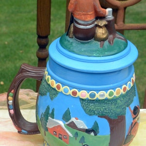 May include: A blue and brown ceramic beer stein with a painted scene of a house and trees. The lid features a figurine of a man in a brown hat and orange jacket.