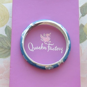 May include: A blue and silver bangle bracelet with a white duck and the words "Quackers Factory" on a pink background.