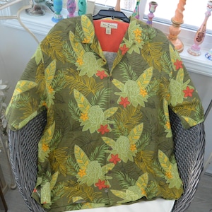 K&ouml;nnte beinhalten: Olivgr&uuml;nes Button-Down-Hemd mit tropischem Blumenmuster. Das Hemd hat gro&szlig;e gr&uuml;ne und gelbe Blumen, rote Akzente und ein hellorangefarbenes Innenfutter. Das Hemd wird auf einem schwarzen Kleiderb&uuml;gel pr&auml;sentiert.