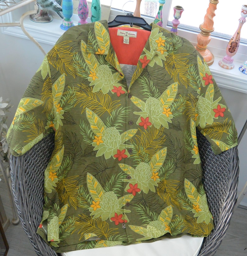 K&ouml;nnte beinhalten: Olivgr&uuml;nes Button-Down-Hemd mit tropischem Blumenmuster. Das Hemd hat gro&szlig;e gr&uuml;ne und gelbe Blumen, rote Akzente und ein hellorangefarbenes Innenfutter. Das Hemd wird auf einem schwarzen Kleiderb&uuml;gel pr&auml;sentiert.