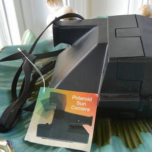May include: A black Polaroid Sun Camera with a black strap and a product tag. The camera is on a teal-colored decorative plate. The tag has the words "Polaroid Sun Camera".