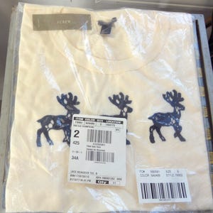 Peut inclure: Un t-shirt à manches courtes de couleur crème orné de trois appliques de rennes en dentelle bleu foncé sur le devant. Le vêtement est emballé dans un sac en plastique transparent avec une étiquette J.Crew et une étiquette blanche avec codes-barres.