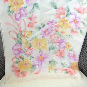 May include: A cream-colored scarf with a floral pattern featuring pink, yellow, and purple flowers, accented with green leaves and pink ribbons. The scarf is made of a soft, lightweight material.