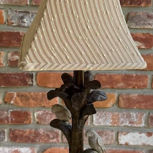 May include: A decorative table lamp with a brown base and a palm tree design. The lamp features two small bird figurines and a beige lampshade with vertical stripes. The lamp is on a wooden surface.