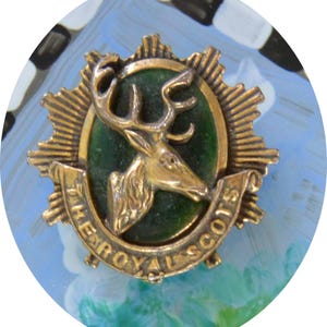 May include: A gold-colored pin featuring a stag's head on a green oval, surrounded by a sunburst design. The text "THE ROYAL SCOTS" is inscribed on a banner below the stag. The pin is set against a blue and white background.