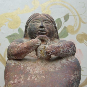 May include: A terracotta sculpture of a seated woman with a round face and a simple hairstyle. The sculpture is weathered and has a reddish-brown colour.