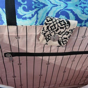 May include: A tote bag with a pink interior featuring black vertical stripes and small heart-shaped designs. A small, patterned wallet with a silver clasp is attached. The background includes a blue patterned fabric.