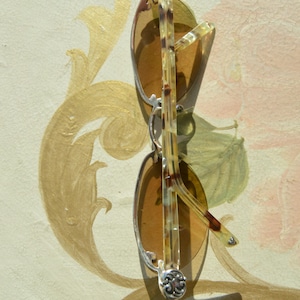 May include: Sunglasses with brown tinted lenses and silver frames. The temples are a light brown and dark brown pattern. The hinges have a decorative silver design. The sunglasses are resting on a painted surface.