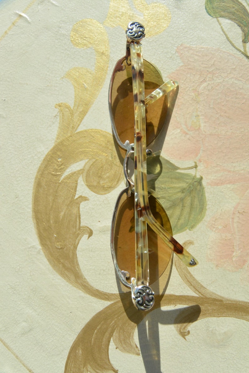 May include: Sunglasses with brown tinted lenses and silver frames. The temples are a light brown and dark brown pattern. The hinges have a decorative silver design. The sunglasses are resting on a painted surface.