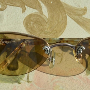 May include: Oval-shaped sunglasses with a light brown tint. The frame is silver-colored with decorative accents on the temples. The brand name "Brighton" is visible on the left lens.