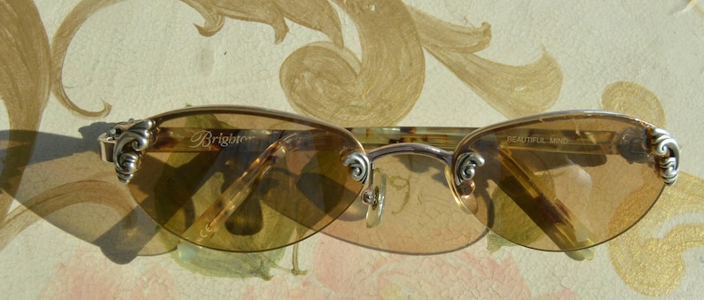 May include: Oval-shaped sunglasses with a light brown tint. The frame is silver-colored with decorative accents on the temples. The brand name "Brighton" is visible on the left lens.
