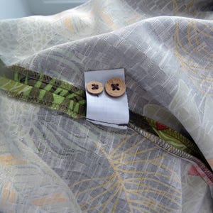 K&ouml;nnte beinhalten: Nahaufnahme eines Stoffes mit grauem und wei&szlig;em Blumenmuster. Zwei Holzkn&ouml;pfe sind an einem wei&szlig;en Etikett befestigt. Der Stoff hat einen gr&uuml;n-braunen Rand mit floralem Design.