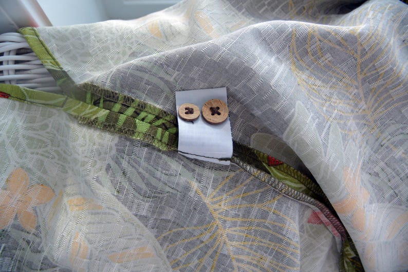 K&ouml;nnte beinhalten: Nahaufnahme eines Stoffes mit grauem und wei&szlig;em Blumenmuster. Zwei Holzkn&ouml;pfe sind an einem wei&szlig;en Etikett befestigt. Der Stoff hat einen gr&uuml;n-braunen Rand mit floralem Design.