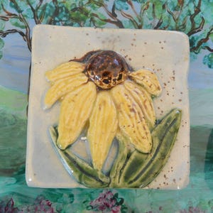 May include: A square ceramic tile featuring a raised, three-dimensional yellow flower with a dark brown center and green leaves. The tile has a light blue-grey background with small brown speckles, showcasing a glossy, hand-glazed finish.