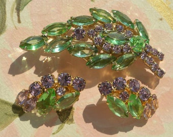 Peut inclure: Un ensemble de bijoux doré composé d'une grande broche en forme de feuille et de deux boucles d'oreilles à clip assorties. Les pièces présentent des cristaux vert clair taille marquise et des strass ronds lavande.