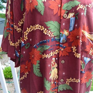 May include: Burgundy button-down shirt with a tropical print. The design includes red poinsettias, green leaves, yellow flowers, palm trees, and a woman in a Santa hat. The shirt has the text "Tommy Bahama" and "Every Day is a Wave".