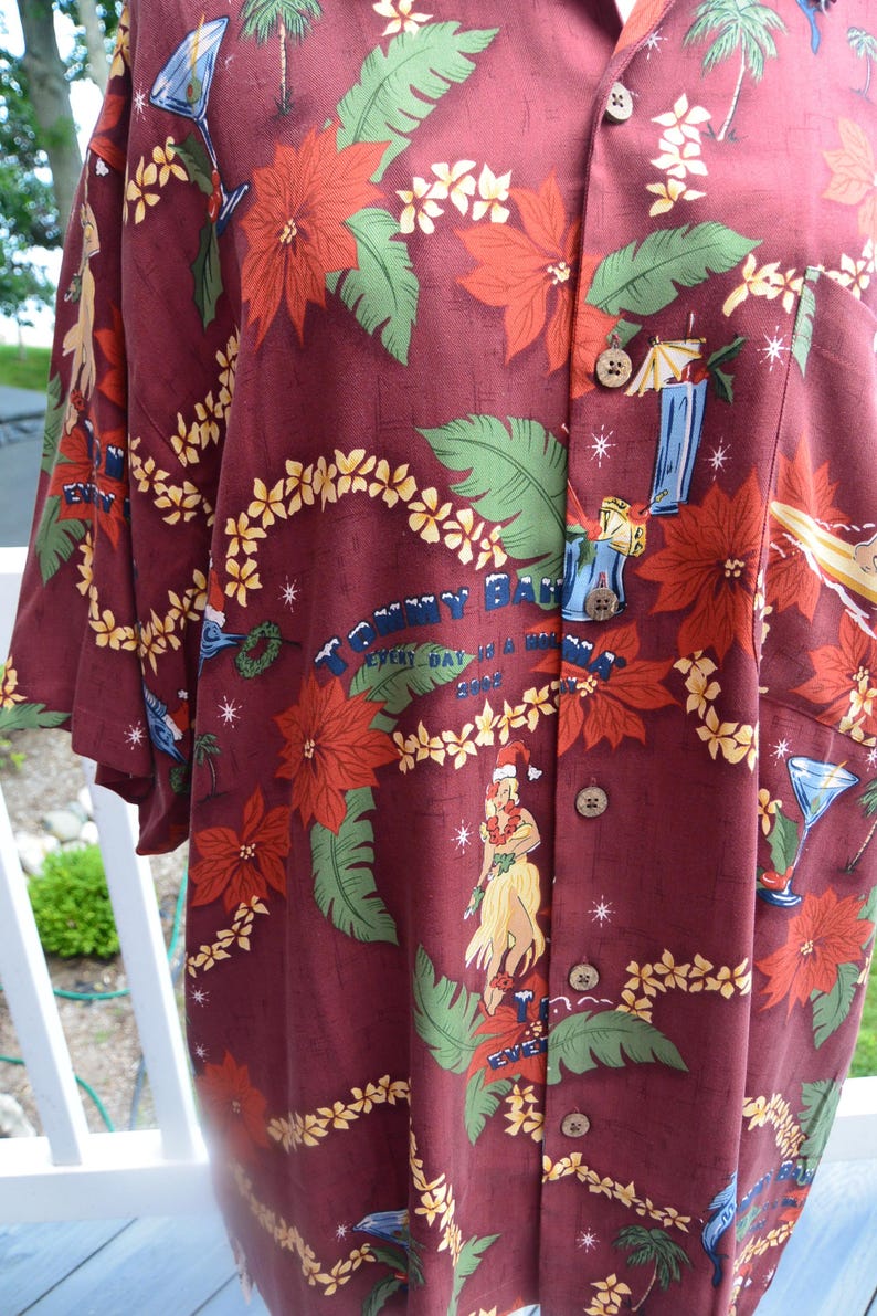 May include: Burgundy button-down shirt with a tropical print. The design includes red poinsettias, green leaves, yellow flowers, palm trees, and a woman in a Santa hat. The shirt has the text "Tommy Bahama" and "Every Day is a Wave".