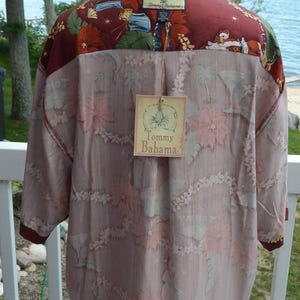 May include: A button-down shirt with a floral pattern in shades of pink and white, with a burgundy collar and cuffs. The shirt has a tag that reads "Tommy Bahama". The shirt is displayed on a white mannequin head.