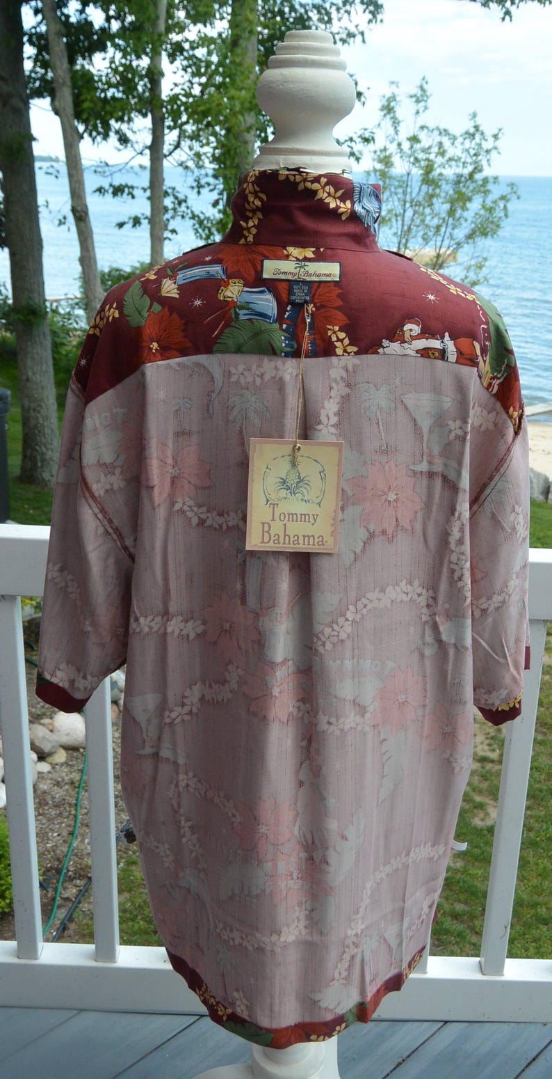 May include: A button-down shirt with a floral pattern in shades of pink and white, with a burgundy collar and cuffs. The shirt has a tag that reads "Tommy Bahama". The shirt is displayed on a white mannequin head.