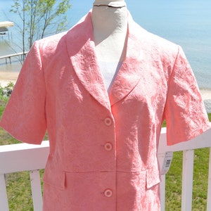 Op de afbeelding: Een koraalkleurige blazer met korte mouwen, met een subtiel bloemenpatroon. De jas heeft een reverskraag, drie knopen en twee voorzakken. De blazer wordt buiten op een mannequin getoond.