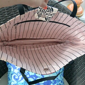 May include: An open tote bag with black handles and a black zip. The interior is lined with pink fabric, featuring black vertical stripes and small black icons. The bag's flap has a black and white pattern.