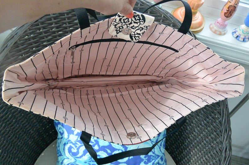 May include: An open tote bag with black handles and a black zip. The interior is lined with pink fabric, featuring black vertical stripes and small black icons. The bag's flap has a black and white pattern.