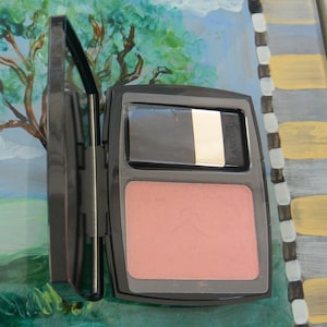 May include: A black compact with a blush and a mirror. The blush is a light coral color. The compact is open, revealing the blush and a mirror with a black, white, and gold accent. The brand name "Lancome" is visible on the mirror.
