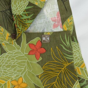 K&ouml;nnte beinhalten: Nahaufnahme eines gr&uuml;nen Stoffes mit tropischem Blumenmuster. Das Design zeigt gro&szlig;e gr&uuml;ne Bl&auml;tter, gelbe und orangefarbene Bl&uuml;ten und ein helles Innenfutter. Ein kleines braunes Etikett mit Logo ist sichtbar.