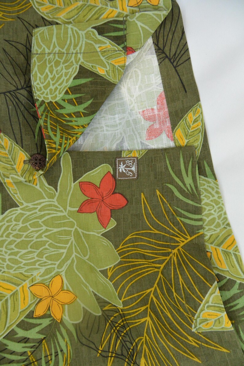 K&ouml;nnte beinhalten: Nahaufnahme eines gr&uuml;nen Stoffes mit tropischem Blumenmuster. Das Design zeigt gro&szlig;e gr&uuml;ne Bl&auml;tter, gelbe und orangefarbene Bl&uuml;ten und ein helles Innenfutter. Ein kleines braunes Etikett mit Logo ist sichtbar.