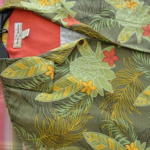 K&ouml;nnte beinhalten: Olivgr&uuml;nes Hemd mit tropischem Blumenmuster in Gelb, Orange und Hellgr&uuml;n. Das Hemd hat eine Brusttasche und ein rotes Innenfutter. Das Markenetikett ist sichtbar.