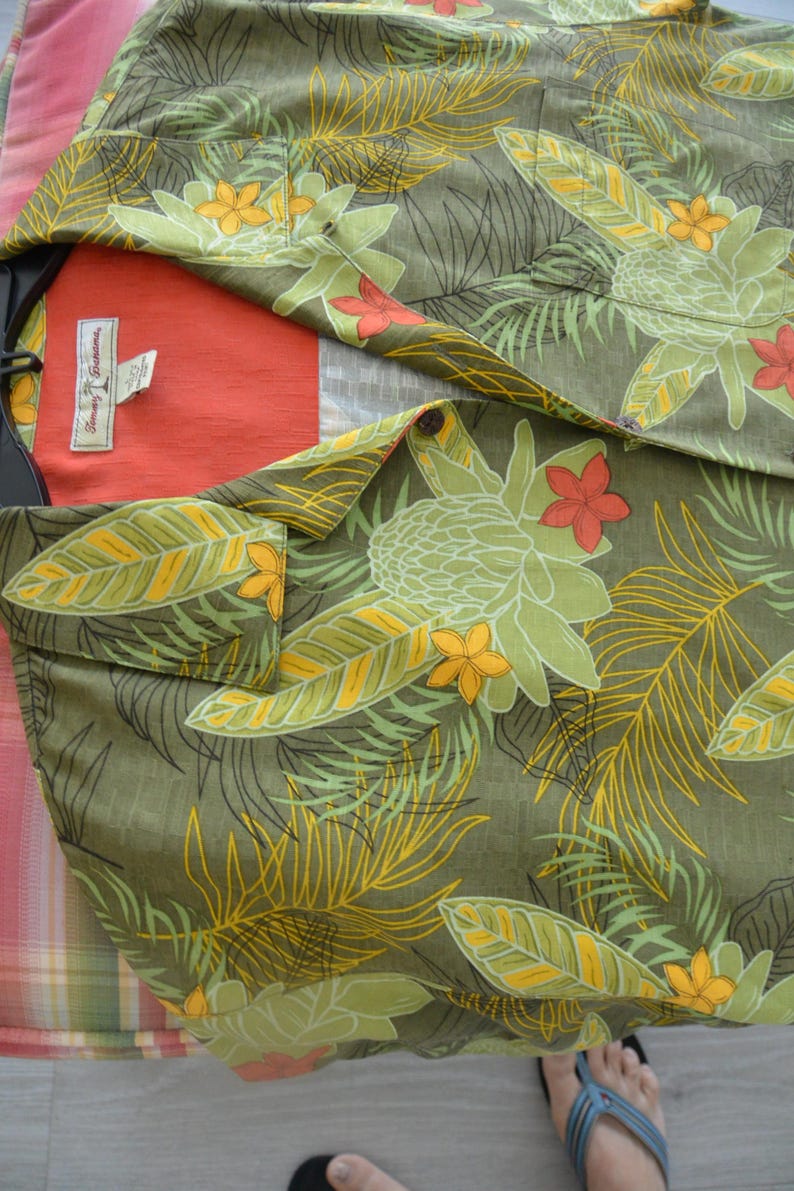 K&ouml;nnte beinhalten: Olivgr&uuml;nes Hemd mit tropischem Blumenmuster in Gelb, Orange und Hellgr&uuml;n. Das Hemd hat eine Brusttasche und ein rotes Innenfutter. Das Markenetikett ist sichtbar.