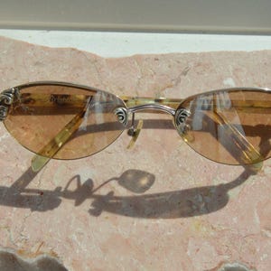 May include: Oval-shaped sunglasses with a gold-colored frame and tinted lenses. The temples feature decorative silver accents. The sunglasses are resting on a pinkish-tan surface.